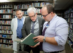 Donn Dexter, left, and Mayo library supervisor Matthew Hoy, right, share vintage newspaper articles, artwork and postcards of Midelfort history with Kristoffer Hellum.